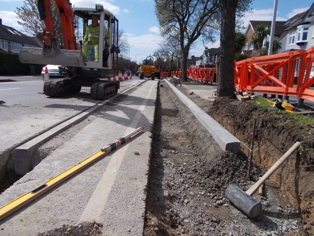 The view along a street with a close up of a new kerb line being laid on concrete. There is the old road to the right on which an orange-boomed mini-excavator sits. To the left there is a gradd verge containing trees.